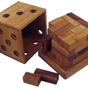 May include: A wooden puzzle cube with a dice-shaped outer frame and a separate, interlocking puzzle piece. The cube is made of light brown wood, with circular holes representing dice pips. Additional wooden pieces are nearby.