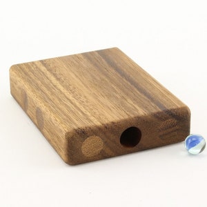 May include: A rectangular wooden box with rounded edges, featuring a circular hole and three decorative circular inlays. A small blue and white marble rests nearby. The box is made of a light brown wood.