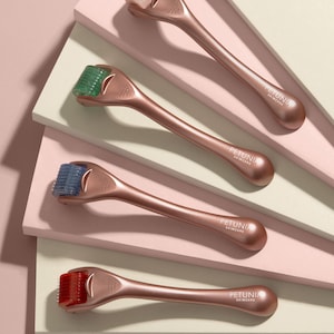 Puede incluir: Cuatro rodillos de derma de oro rosa con agujas de diferentes colores. Los rodillos están dispuestos en forma de abanico sobre un fondo rosa y beige. El nombre de la marca "PETUNIA SKINCARE" está impreso en cada rodillo.
