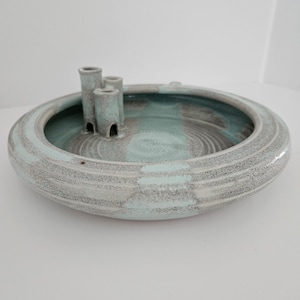 May include: A round, ceramic dish with a light blue and gray glaze. The dish has a raised center with three small, cylindrical openings.