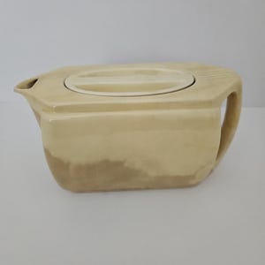 May include: A cream-colored ceramic teapot with a rectangular body and a matching lid. The teapot has a handle on one side and a spout on the other. The bottom of the teapot has a darker, mottled appearance.