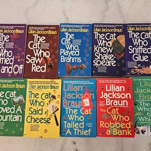 May include: A collection of vintage paperback books by Lilian Jackson Braun, featuring the "Cat Who" series. The covers display titles like "The Cat Who Turned On and Off," and "The Cat Who Saw Red," with colorful designs and cat illustrations.