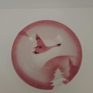 May include: A small, round, pink and white bowl with a painted design. The design features a flying bird, two stylized evergreen trees, and a pink border. The bowl is likely made of ceramic or glass.
