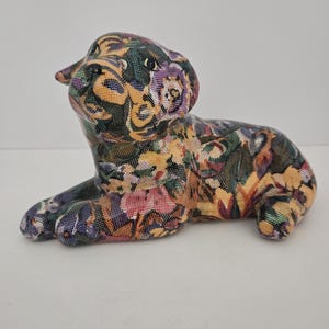 May include: A ceramic dog figurine in a reclining pose, adorned with a floral pattern in shades of yellow, purple, green, and pink. The dog is lying down with its head raised, showcasing the intricate design and craftsmanship of the decorative piece.