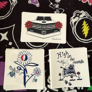 May include: Three white stickers with unique designs are displayed on a black patterned surface. One sticker features a car design with red flowers, another shows a flower with bees, and the last one has the text "High on the mountain".