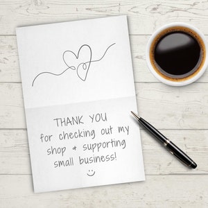 K&ouml;nnte beinhalten: Eine wei&szlig;e Karte mit einer handgeschriebenen Nachricht, die "THANK YOU for checking out my shop & supporting small business!" mit einem Smiley am unteren Rand lautet. Ein schwarzer Stift und eine wei&szlig;e Kaffeetasse befinden sich auf einer wei&szlig;en Holzoberfl&auml;che.