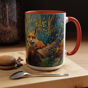 May include: A white ceramic mug with a red rim and handle. The mug features a colorful illustration of a fox sitting on a log in a forest setting. The background includes trees, leaves, and flowers.