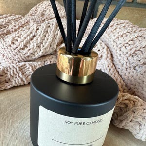Peut inclure: Un diffuseur noir avec un bord dor&eacute; et des b&acirc;tonnets noirs. L'&eacute;tiquette indique "SOY PURE CANDLES". L'arri&egrave;re-plan comprend une couverture beige tricot&eacute;e.