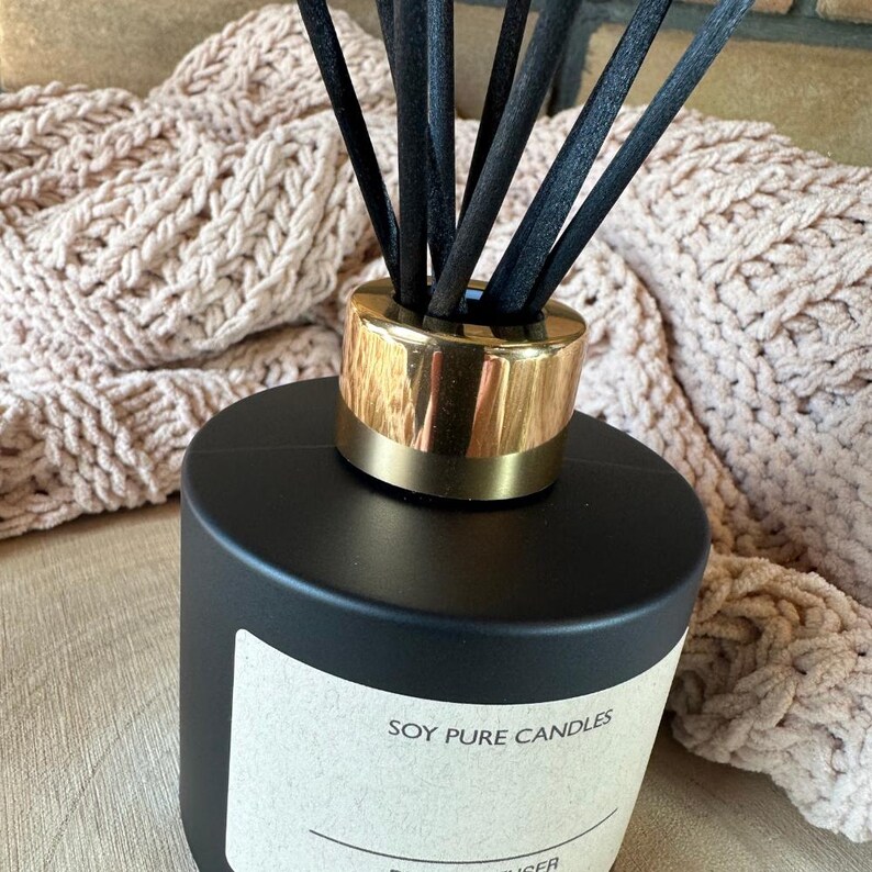 Peut inclure: Un diffuseur noir avec un bord dor&eacute; et des b&acirc;tonnets noirs. L'&eacute;tiquette indique "SOY PURE CANDLES". L'arri&egrave;re-plan comprend une couverture beige tricot&eacute;e.