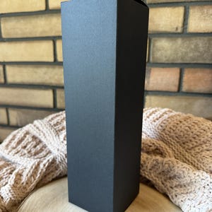 Peut inclure: Une bo&icirc;te cadeau noire, rectangulaire et haute, avec une surface textur&eacute;e. La bo&icirc;te est pos&eacute;e verticalement sur une surface en bois, avec une couverture en tricot beige en arri&egrave;re-plan. La bo&icirc;te est vide et pr&ecirc;te &agrave; &ecirc;tre remplie.