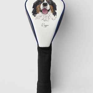 May include: White golf club head cover with a black and white Bernese Mountain Dog illustration and the name "Reggie" printed on it. The cover has a blue trim and a black knitted shaft.