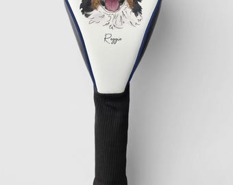Op de afbeelding: Witte golfclubkophoes met een zwart-witte illustratie van een Berner Sennenhond en de naam "Reggie" erop gedrukt. De hoes heeft een blauwe rand en een zwarte gebreide schacht.