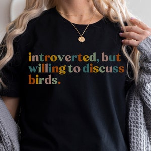 Op de afbeelding: Een zwart T-shirt met de tekst "introvert, maar bereid om over vogels te praten." in kleurrijke letters.