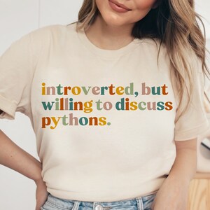 May include: A cream-colored t-shirt with the text "introverted, but willing to discuss pythons" in colorful, retro-style lettering. The shirt has short sleeves and a crew neck.