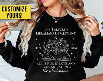May include: Black sweatshirt with white text that reads "The Tortured Librarians Department Est 2024 All is fair in love and classification Mrs. Johnson" with a white outline of a book and flowers.