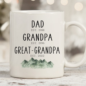 K&ouml;nnte beinhalten: Wei&szlig;e Keramiktasse mit schwarzem Text, der "Dad Est. 1988 Grandpa Est. 1996 Great-Grandpa Est. 2022" lautet, mit einer gr&uuml;nen Aquarell-Bergkette am Boden.