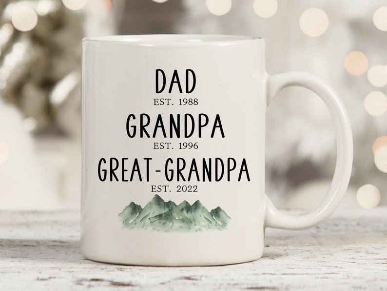 K&ouml;nnte beinhalten: Wei&szlig;e Keramiktasse mit schwarzem Text, der "Dad Est. 1988 Grandpa Est. 1996 Great-Grandpa Est. 2022" lautet, mit einer gr&uuml;nen Aquarell-Bergkette am Boden.