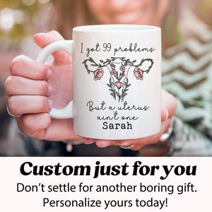 Pu&ograve; includere: Una tazza in ceramica bianca con una grafica nera e rosa di un utero con fiori e il testo "I got 99 problems but a uterus ain't one Sarah".