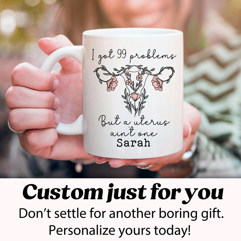 Pu&ograve; includere: Una tazza in ceramica bianca con una grafica nera e rosa di un utero con fiori e il testo "I got 99 problems but a uterus ain't one Sarah".