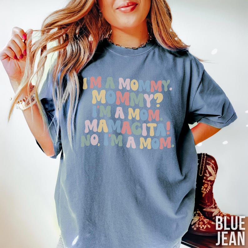 K&ouml;nnte beinhalten: Blaues Jeans-T-Shirt mit dem Text "I'M A MOMMY. MOMMY? I'M A MOM. MAMACITA! NO, I'M A MOM." in farbenfrohen Buchstaben im Retro-Stil. Das Shirt hat eine lockere Passform und kurze &Auml;rmel.
