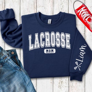 Op de afbeelding: Navyblauwe trui met de tekst "Lacrosse Mom" en een lacrosse-stick afbeelding op de mouw. De mouw heeft ook de naam "Liam" erop.