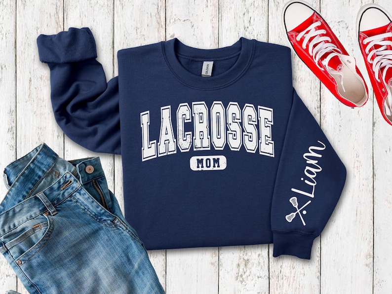 Op de afbeelding: Navyblauwe trui met de tekst "Lacrosse Mom" en een lacrosse-stick afbeelding op de mouw. De mouw heeft ook de naam "Liam" erop.