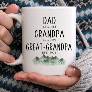 K&ouml;nnte beinhalten: Wei&szlig;e Keramiktasse mit schwarzem Text, der "Dad Est. 1988 Grandpa Est. 1996 Great-Grandpa Est. 2022" lautet, mit einer Aquarell-Illustration von gr&uuml;nen Bergen am Boden.
