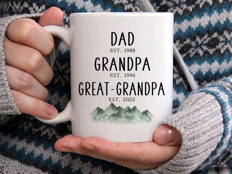 K&ouml;nnte beinhalten: Wei&szlig;e Keramiktasse mit schwarzem Text, der "Dad Est. 1988 Grandpa Est. 1996 Great-Grandpa Est. 2022" lautet, mit einer Aquarell-Illustration von gr&uuml;nen Bergen am Boden.