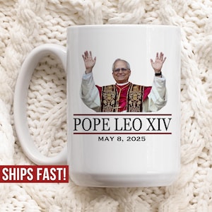 May include: White ceramic mug featuring an image of Pope Leo XIV with his arms raised. The mug has the text "POPE LEO XIV" and "MAY 8, 2025" in black and red. The mug has a white handle.