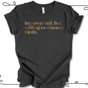 Op de afbeelding: Een donkergrijs T-shirt met de tekst "introvert, maar bereid om over vogels te praten." in kleurrijke letters.
