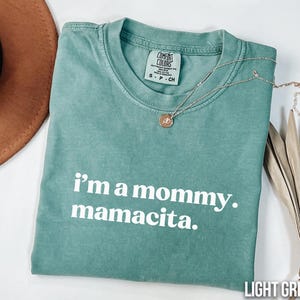 May include: Light green Comfort Colors t-shirt with the text "i'm a mommy. mamacita." in white. A silver necklace with a pendant rests on the shirt. A brown hat and dried leaves are in the background.