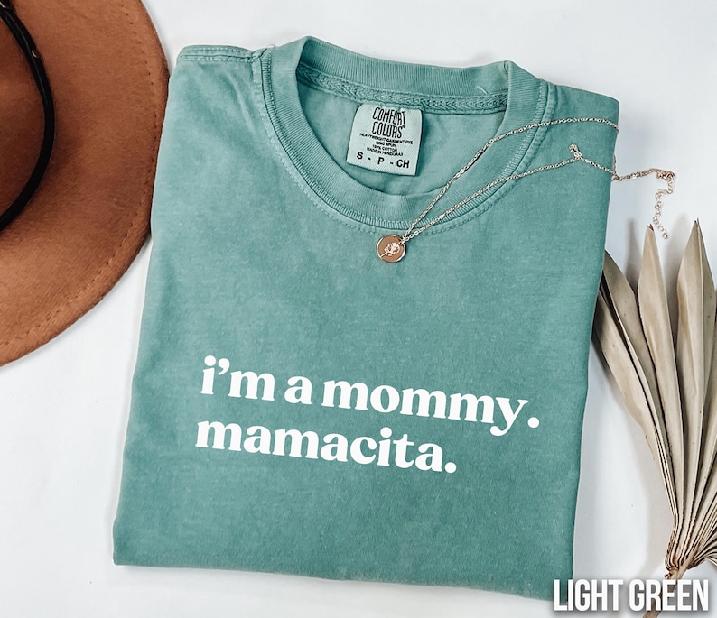 May include: Light green Comfort Colors t-shirt with the text "i'm a mommy. mamacita." in white. A silver necklace with a pendant rests on the shirt. A brown hat and dried leaves are in the background.