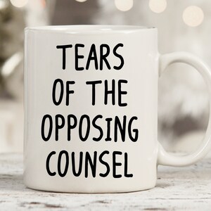 May include: White ceramic coffee mug with black text that reads "Tears of the Opposing Counsel".