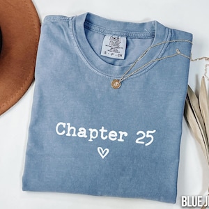 May include: A blue jean colored t-shirt with the words "Chapter 25" and a heart symbol printed in white. A gold necklace with a pendant is draped across the shirt. A brown hat and dried leaves are in the background.