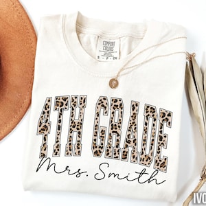 May include: Ivory-colored t-shirt with the words "4TH GRADE" in a leopard print design. Below the text is the name "Mrs. Smith" in a cursive font. The shirt has a round neckline and short sleeves.