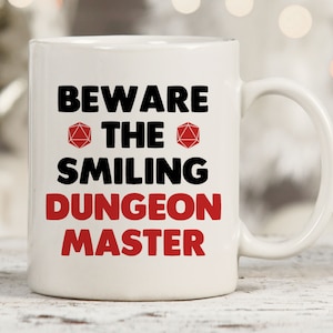 May include: White ceramic coffee mug with a black and red design. The mug reads "Beware the Smiling Dungeon Master" with a red 20-sided die on either side of the word "the".
