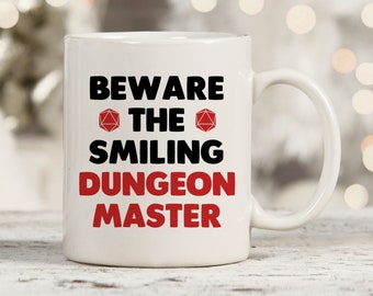 May include: White ceramic coffee mug with a black and red design. The mug reads "Beware the Smiling Dungeon Master" with a red 20-sided die on either side of the word "the".