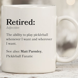 May include: White ceramic coffee mug with a black text design that defines "Retired" as the ability to play pickleball whenever and wherever you want. The text also includes "See also: Matt Parmley, Pickleball Fanatic."