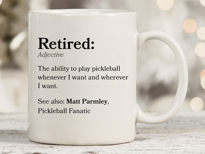 May include: White ceramic coffee mug with a black text design that defines "Retired" as the ability to play pickleball whenever and wherever you want. The text also includes "See also: Matt Parmley, Pickleball Fanatic."