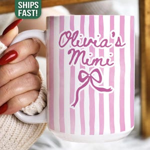 Könnte beinhalten: Weißer Keramikbecher mit rosa vertikalen Streifen und dem Text "Olivia's Mimi" in Schreibschrift, zusammen mit einer Schleifengrafik. Der Becher hat einen weißen Griff.