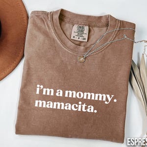 May include: Espresso-coloured t-shirt with the text "i'm a mommy. mamacita." in white. The shirt has a crew neck and short sleeves. A silver necklace with a pendant is draped across the shirt. A brown hat and dried leaves are in the background.