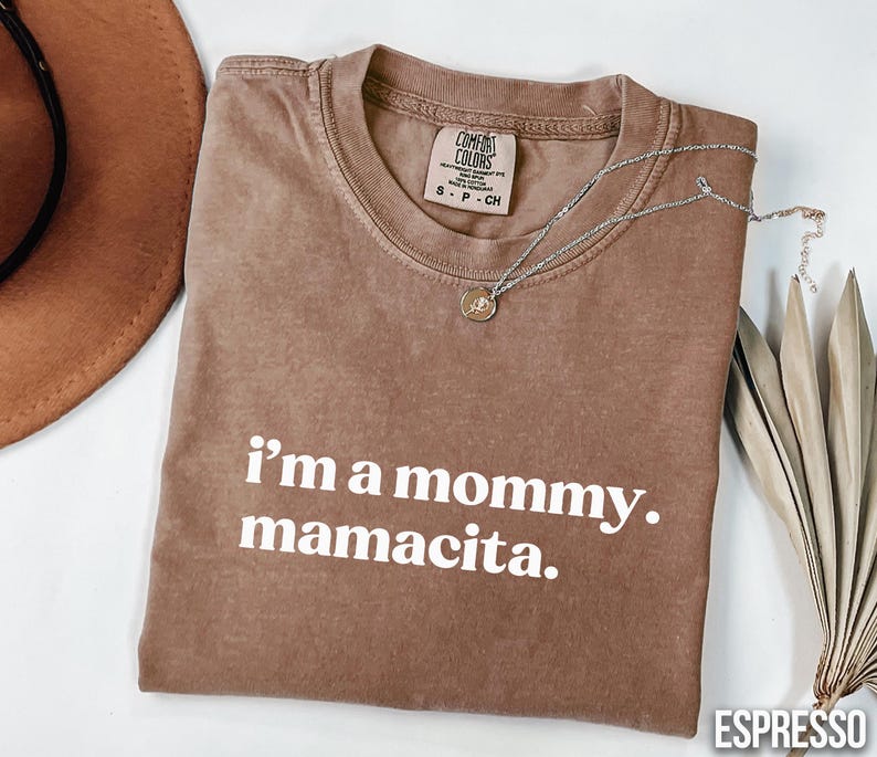 May include: Espresso-coloured t-shirt with the text "i'm a mommy. mamacita." in white. The shirt has a crew neck and short sleeves. A silver necklace with a pendant is draped across the shirt. A brown hat and dried leaves are in the background.