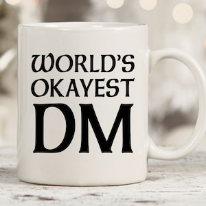 May include: White ceramic mug with black text that reads "WORLD'S OKAYEST DM".