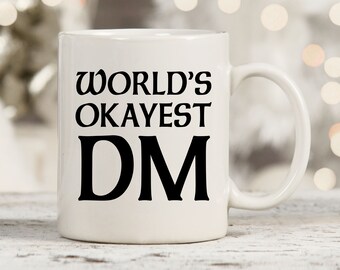 May include: White ceramic mug with black text that reads "WORLD'S OKAYEST DM".