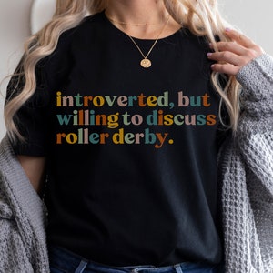 K&ouml;nnte beinhalten: Ein schwarzes T-Shirt mit dem Text "introvertiert, aber bereit, &uuml;ber Roller Derby zu diskutieren." in bunten Buchstaben.