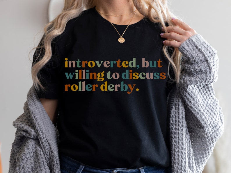 K&ouml;nnte beinhalten: Ein schwarzes T-Shirt mit dem Text "introvertiert, aber bereit, &uuml;ber Roller Derby zu diskutieren." in bunten Buchstaben.