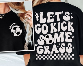 Op de afbeelding: Zwart T-shirt met witte tekst die "Let's Go Kick Some Grass" luidt in een retro lettertype. Een voetbal is te zien in de tekst. Het shirt heeft ook een geruit patroon aan de onderkant.