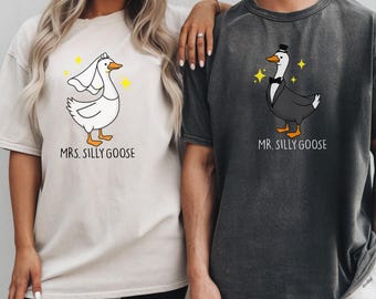 May include: Two matching t-shirts, one beige and one charcoal gray. The beige shirt features a cartoon goose wearing a veil with the text "MRS. SILLY GOOSE". The charcoal shirt has a goose in a tuxedo with the text "MR. SILLY GOOSE".