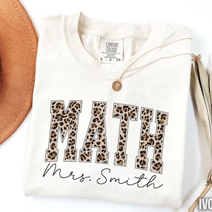 May include: Ivory-colored Comfort Colors t-shirt with the word "MATH" in a leopard print design. Below the word "MATH" is the name "Mrs. Smith" in a handwritten font. A gold necklace and a brown hat are also in the image.