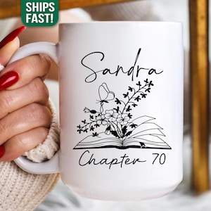 May include: White ceramic mug with a black floral and butterfly design, the name "Sandra" in cursive, and the text "Chapter 70." The mug is held by a person wearing a cream-colored sweater.